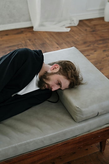 man, black long sleeve shirt, lying, gray couch, indoor, contemplative mood, deep thought, wooden floor, casual attire, relaxed pose, home setting