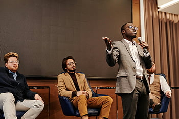 man in gray suit, holding microphone, business panel discussion, diverse professionals, speaker presenting, panel discussion, business event, professional event, conference, presentation, audience engagement