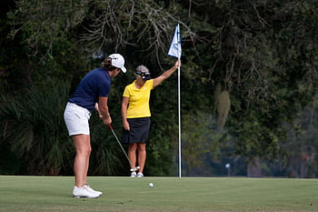 women playing golf, golf course, putting green, golf club, golf flag, outdoor sports, female golfers, golf attire, golfing activity, green grass, trees in background