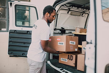 volunteer worker, food labelled boxes, white van, community distribution, aid supplies, cardboard boxes, trunk, loading supplies, food aid, van trunk, distribution