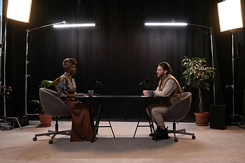 podcast recording, man and woman, studio setup, microphones, studio lights, well-lit studio, hosts, table, chairs, plants, black background