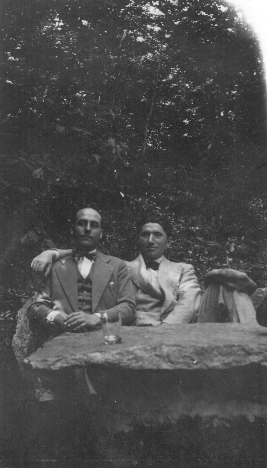 black and white, two men, suits, bow ties, outdoor setting, trees, stone table, vintage photo, casual pose, drinking glass, nature background