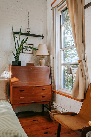old bedroom, vintage wooden desk, plants by window, warm bedroom decor, inviting bedroom, brick wall, wooden floor, cozy interior, natural light, retro furniture, window view