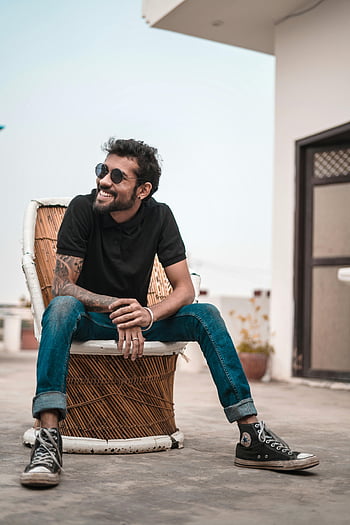 man, black polo shirt, blue faded jeans, sunglasses, tattoos, sitting outdoors, smiling, rooftop, stylish, casual fashion, converse shoes