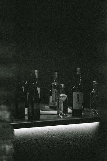 bottles on table, grayscale photography, dimly lit, black and white, bar counter, mysterious atmosphere, various bottles, dark setting, monochrome image, low light, shadowy scene