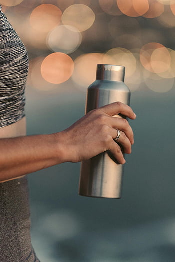 stainless steel vacuum flask, reusable steel water bottle, eco-friendly living, bokeh background, close-up, woman holding, outdoors, sustainable lifestyle, metal flask, blurred background, reusable bottle