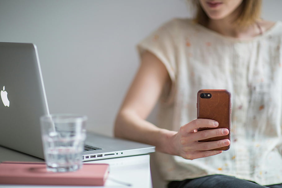 tenendo smartphone, giovane donna, Utilizzo dello smartphone, il computer portatile, scrivania, in casa, bicchiere d'acqua, Abbigliamento casual, focalizzata, tecnologia, spazio di lavoro