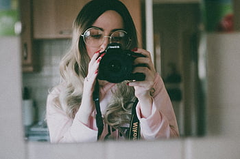 woman holding camera, mirror selfie, professional camera, indoors, young woman, taking photo, indoor photography, mirror reflection, casual outfit, glasses, blonde hair