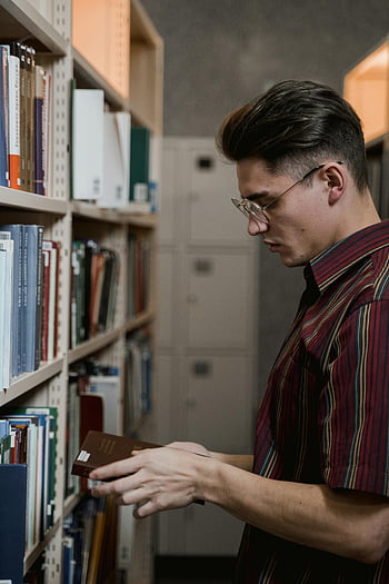 man in eyeglasses, holding a book, library, browsing bookshelf, selecting book, man reading, eyeglasses, bookshelf, library interior, book selection, man in library