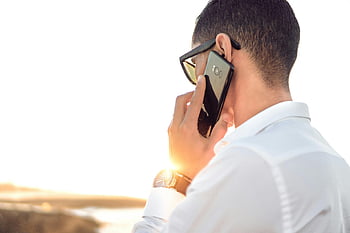 man talking on phone, white collared shirt, black Android smartphone, sunglasses, outdoors Morocco, sunset backlit, shallow focus photography, smartphone conversation, casual business attire, male professional, sunset scene