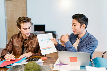 men sitting at table, discussing graphs, data analysis, shared workspace, modern office, two men, professional meeting, business discussion, collaborative work, office setting, modern workspace