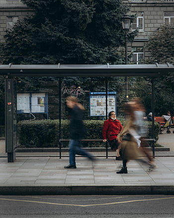 Donna in cappotto rosso, fermata dell'autobus, persone che passano, fermata dell'autobus della città, donna seduta, cappotto rosso, scena urbana, trasporto pubblico, vita di città, fotografia di strada, sfocatura movimento