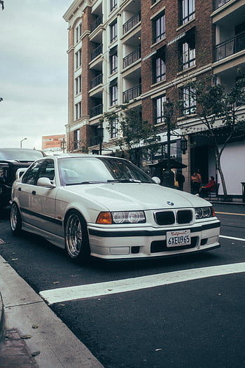 BMW E36, stanced car, white BMW, city street, urban buildings, parked car, E36 coupe, street corner, sleek car, California license plate, modified car