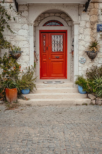 wooden solid house door, rustic entrance, vibrant red door, stone wall, lush plants, arched doorway, potted plants, cobblestone path, outdoor decor, house entrance, red front door