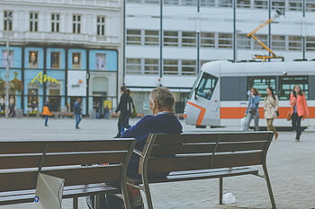elderly man, sitting on bench, city life, urban scene, trams passing, public transportation, cityscape, street view, daily life, outdoor setting, people walking