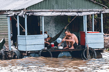 man pouring water, dipper, blue and grey house, flooded wooden houseboat, heavy flood, man sitting, water pouring, flood scene, houseboat living, flood waters, man on houseboat