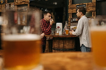 men, bar, beer glasses, rustic setting, indoor bar, chatting, two men, barrel table, casual conversation, wooden decor, beer culture