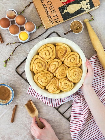 flat-lay photography, person holding bowl, homemade cinnamon rolls, baking process, ingredients on table, top view, round white bowl, baking ingredients, cinnamon rolls recipe, baking tools, flat lay food photography