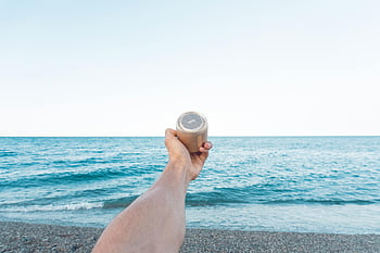 beach, summer vibes, hand holding speaker, brown speaker, sea backdrop, beach leisure, upward speaker, calm sea, summer beach, outdoor speaker, seaside relaxation