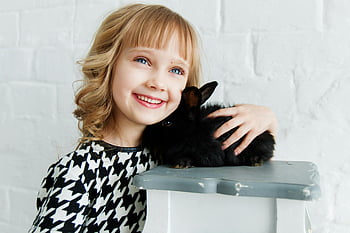 girl holding black rabbit, smiling young girl, blond hair, cute black rabbit, indoors, cuddling rabbit, white brick wall, happy child, pet rabbit, indoor photo, black and white dress