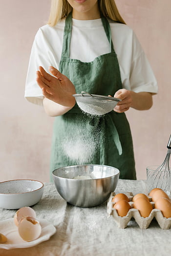 Frau, die Mehl sitzt, grüne Schürze, Backen, Küche, Eier, Sieb, Schüssel, Mehl sieben, Backzutaten, Küchenaktivität, Frau Backen