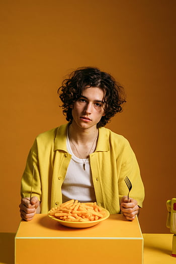 young man, sad face, sitting by table, food, curly hair, eating fries, vibrant retro orange background, yellow jacket, white shirt, plate of fries, orange table